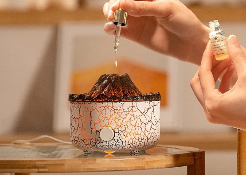 A hand adding a few drops of essential oil from a dropper into the white crackle volcano flame diffuser, small oil bottle held beside it, on a side table in a warm-lit room.