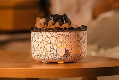White crackle lava-rock volcano flame diffuser on a wood table, a warm orange flame glow of backlit mist rising from the textured crater, single round button on the front, in a dim cozy room.