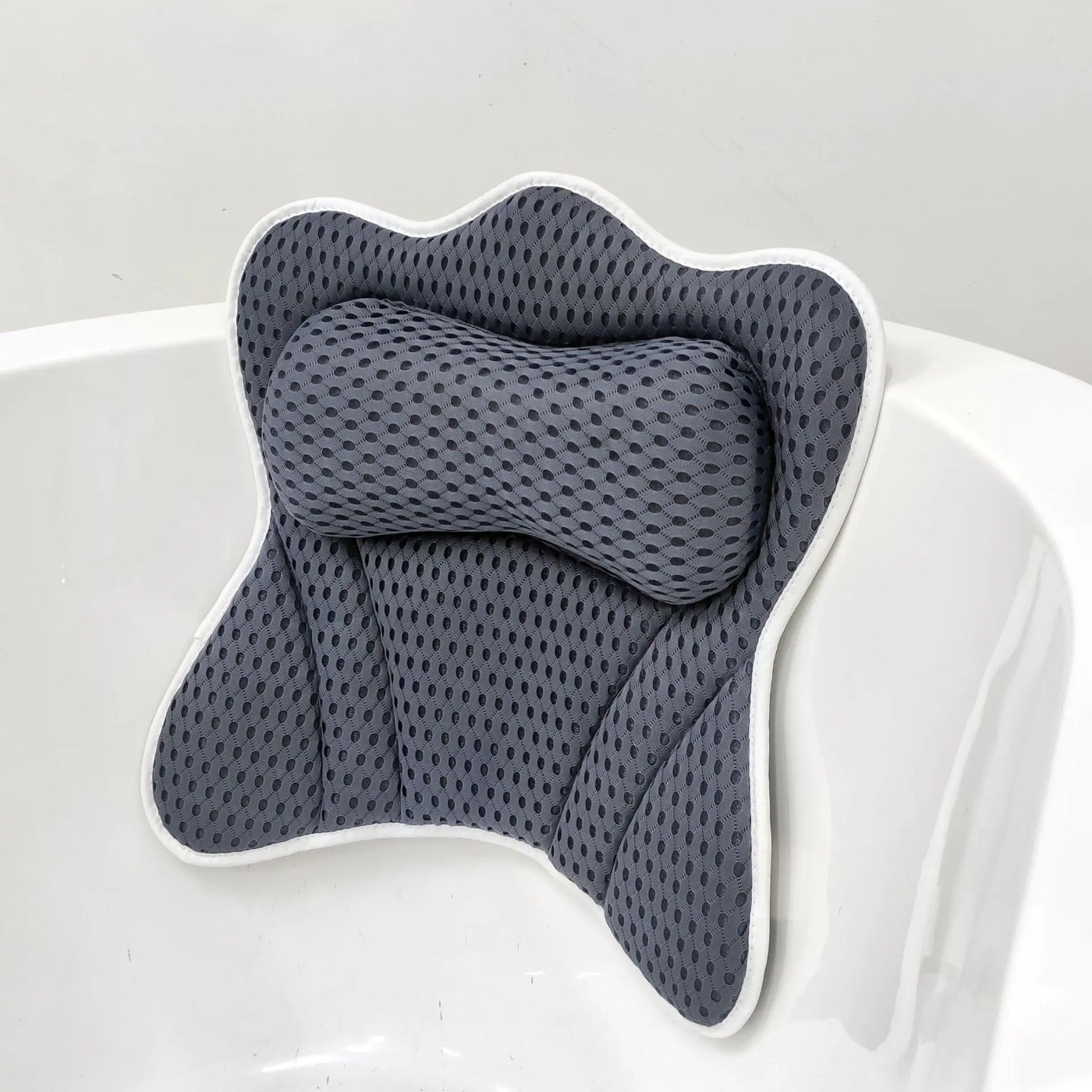 Grey 4D air-mesh bathtub pillow with a raised bone-shaped neck bolster and white piped trim, suction-mounted on the rim of a white bathtub.