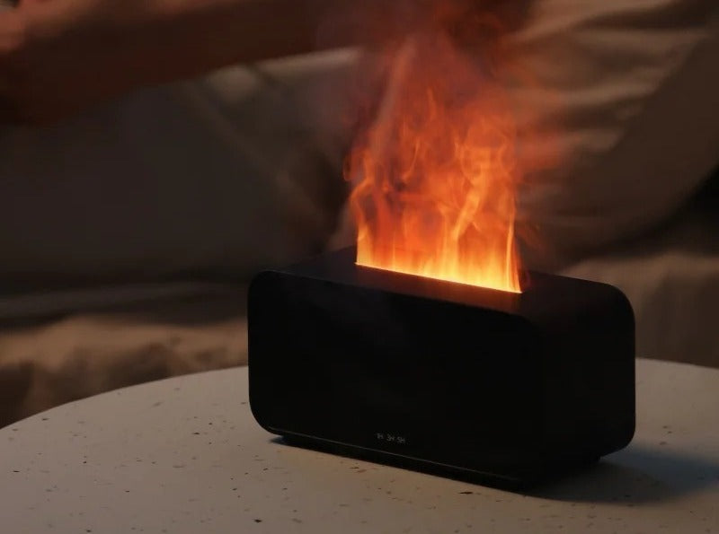 Flame effect diffuser on a bedside table casting a warm flickering flame glow while a person relaxes on the bed behind it — cozy wind-down ambiance.