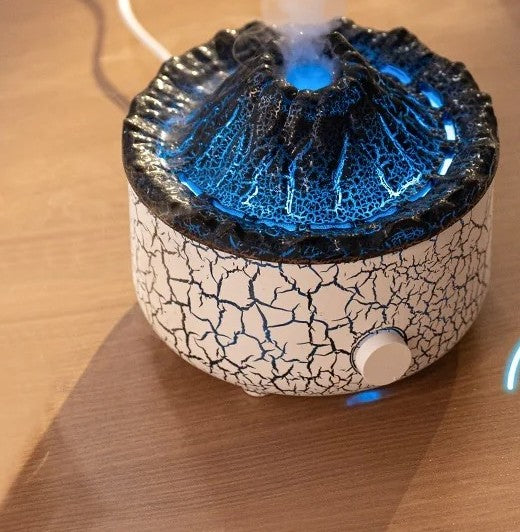 Close-up of the volcano diffuser's textured crater top glowing cool blue with a rising column of cool mist, white crackle-glaze body and front control button visible.